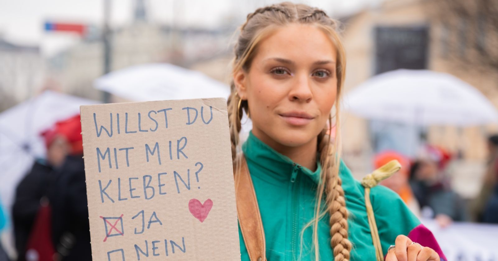 Aktivistin Anja Windl (bekannt als "Klima-Shakira") mit geflochtenen Haaren zeigt bei einer Klimaprotest-Aktion ein Pappschild mit der Aufschrift „Willst du mit mir kleben? – Ja / Nein“ und einem Herzsymbol.