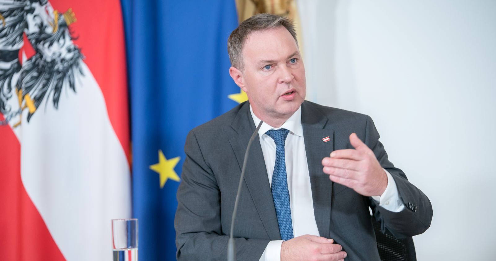 Andreas Babler in grauem Anzug gestikuliert während einer Pressekonferenz, hinter ihm sind die österreichische und die EU-Flagge zu sehen.