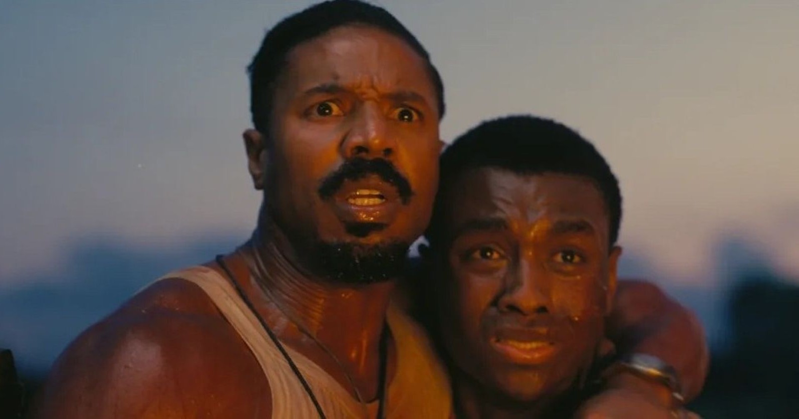 Filmszene aus Blood &amp; Sinners, Zwei Männer mit schweißbedeckten Gesichtern schauen erschrocken in die Ferne, während die Sonne im Hintergrund untergeht.