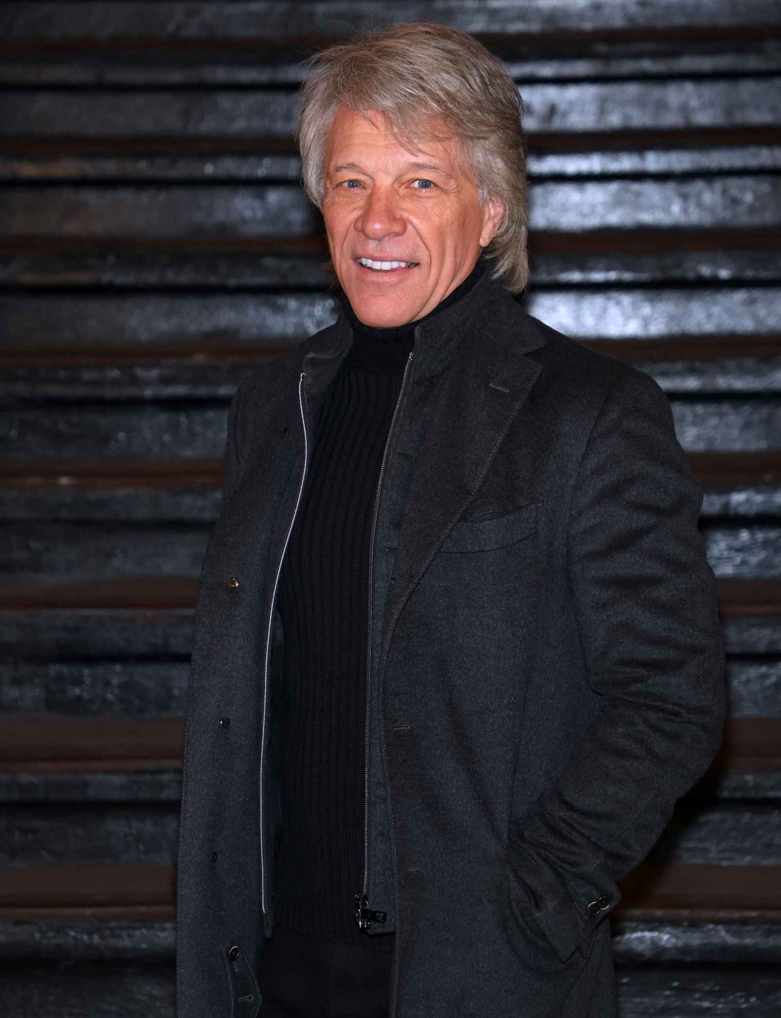 Jon Bon Jovi steht lächelnd vor einer dunklen Treppe und trägt einen dunklen Mantel über einem schwarzen Oberteil.