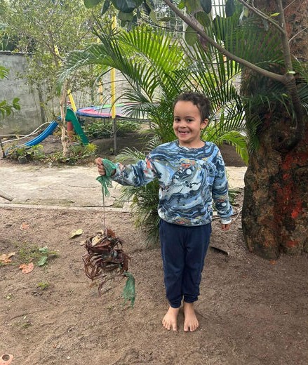 Noah (8) steht barfuß in einem Garten, hält ein gebasteltes Objekt in der Hand; im Hintergrund sind Pflanzen, eine Rutsche und eine Mauer zu sehen.