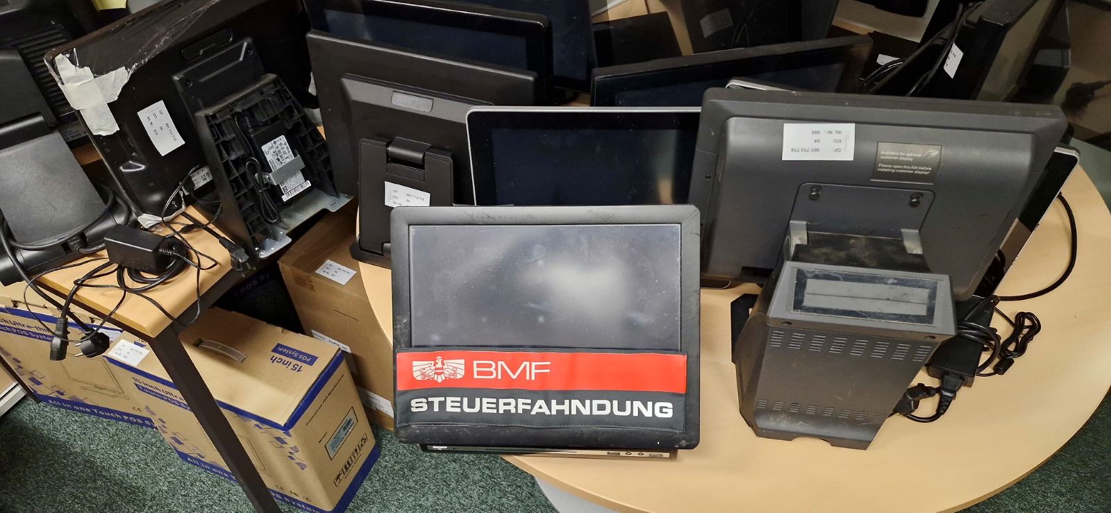 Mehrere beschlagnahmte Registrierkassen und Kassengeräte, darunter ein Monitor mit dem Aufkleber „BMF Steuerfahndung“, auf einem Tisch in einem Ermittlungsraum.