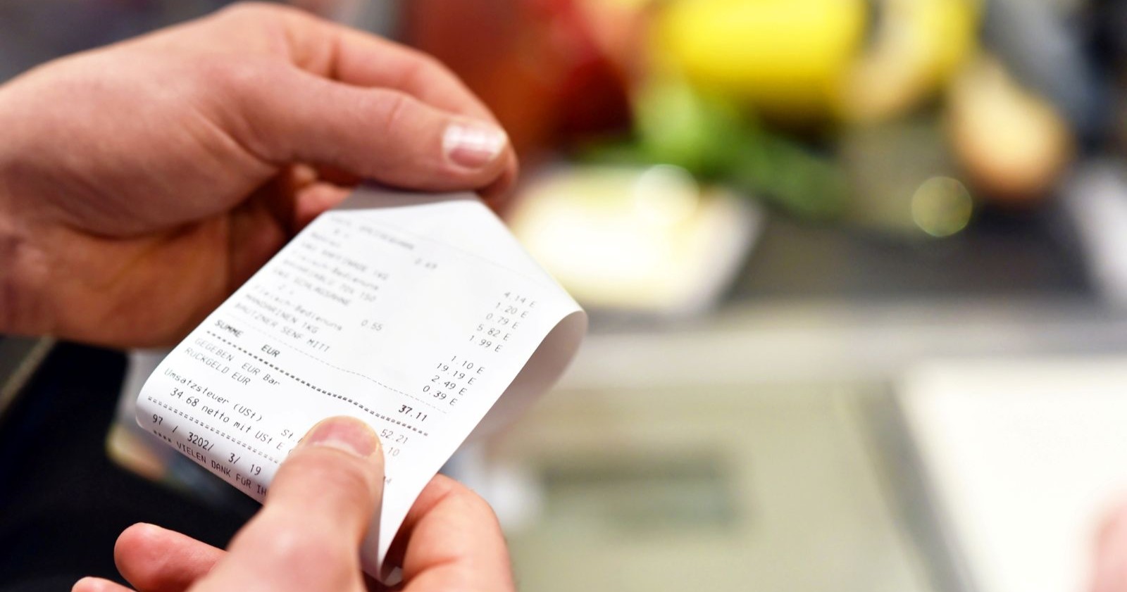 Eine Person hält an der Supermarktkassa einen Kassenbon in der Hand – im Hintergrund sind unscharf Lebensmittel wie Obst und Gemüse zu sehen.