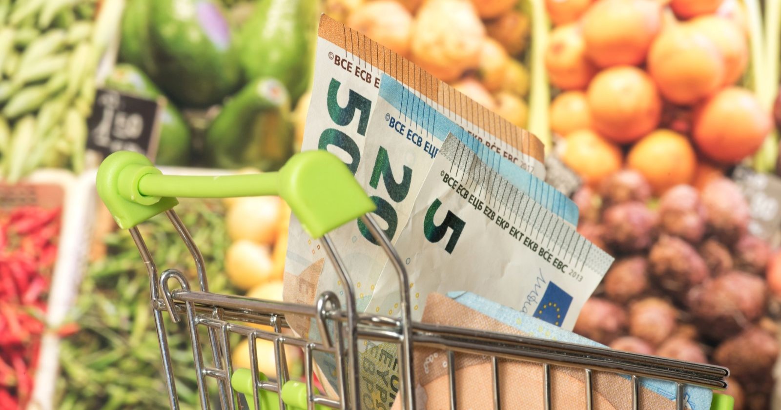 Einkaufswagen mit Euro-Geldscheinen vor einem Gemüseregal im Supermarkt, Symbol für günstigeren Einkauf durch die Mehrwertsteuer-Senkung.