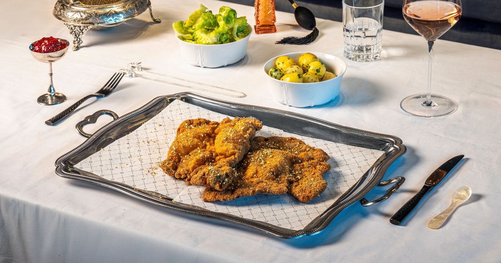 Luxuriös angerichtetes Wagyu-Schnitzel mit Goldflocken auf silberner Platte, dazu Schälchen mit Kaviar, Preiselbeeren, Petersilienkartoffeln und Salat auf weiß gedecktem Tisch.