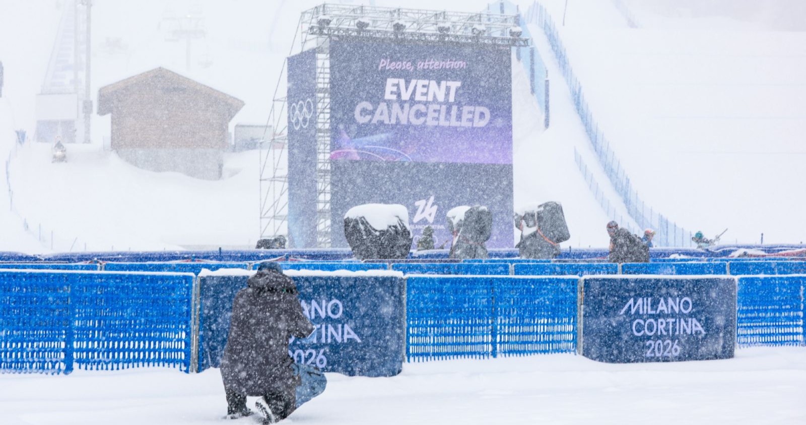 Verschneites Slopestyle-Gelände bei den Olympischen Winterspielen 2026 in Livigno. Auf einer großen Videowand steht „Event Cancelled“, davor Absperrungen mit der Aufschrift „Milano Cortina 2026“, dichter Schneefall und mehrere Personen im Sturm.