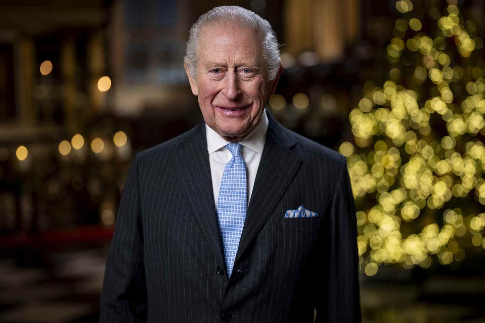 Porträt von König Charles III. im dunklen Anzug mit hellblauer Krawatte vor festlich beleuchtetem Hintergrund.