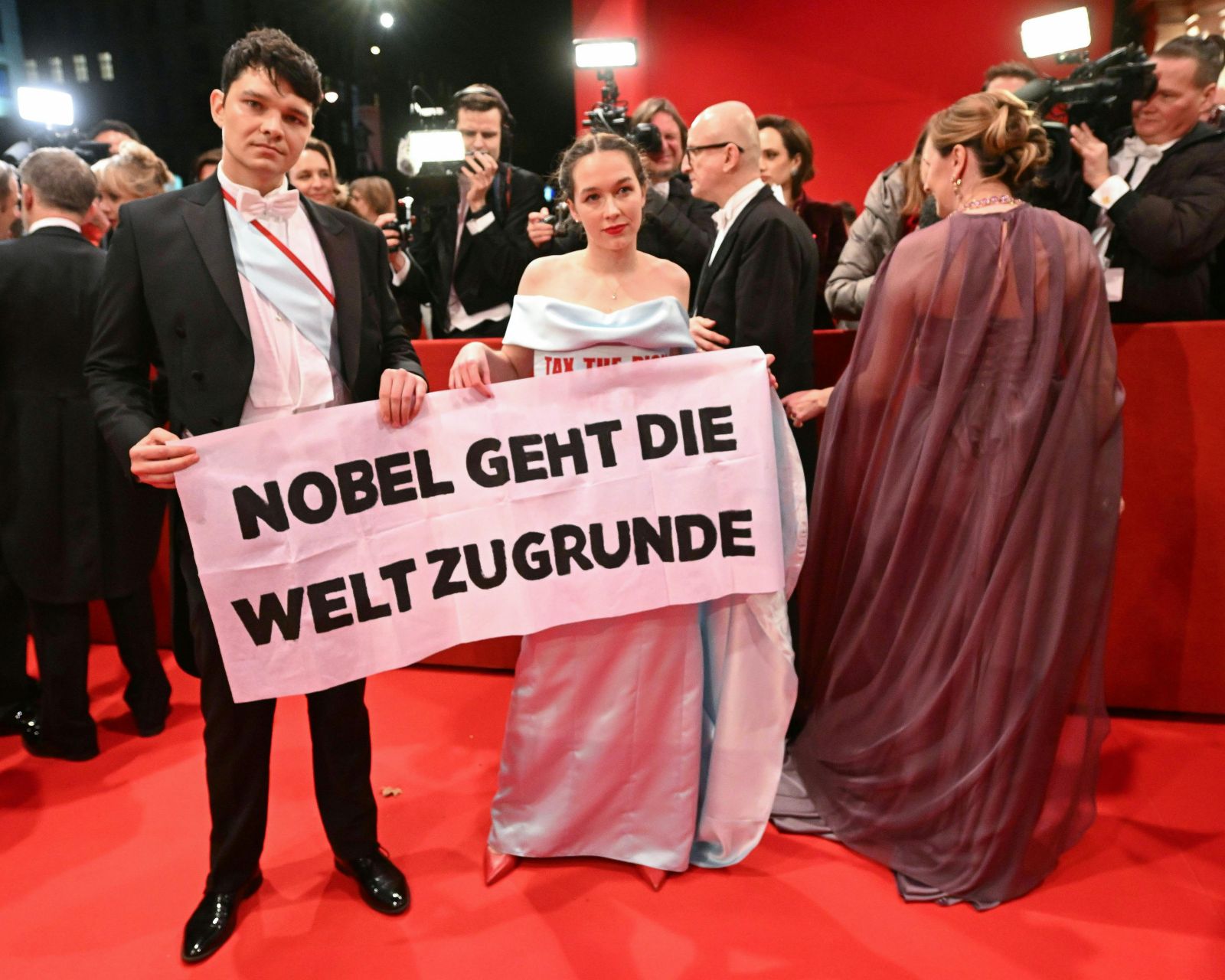 Lena Schilling und ein Begleiter beim Wiener Opernball, sie halten ein Banner mit der Aufschrift „Nobel geht die Welt zugrunde“, umgeben von Fotografen und Kameras auf dem Roten Teppich.