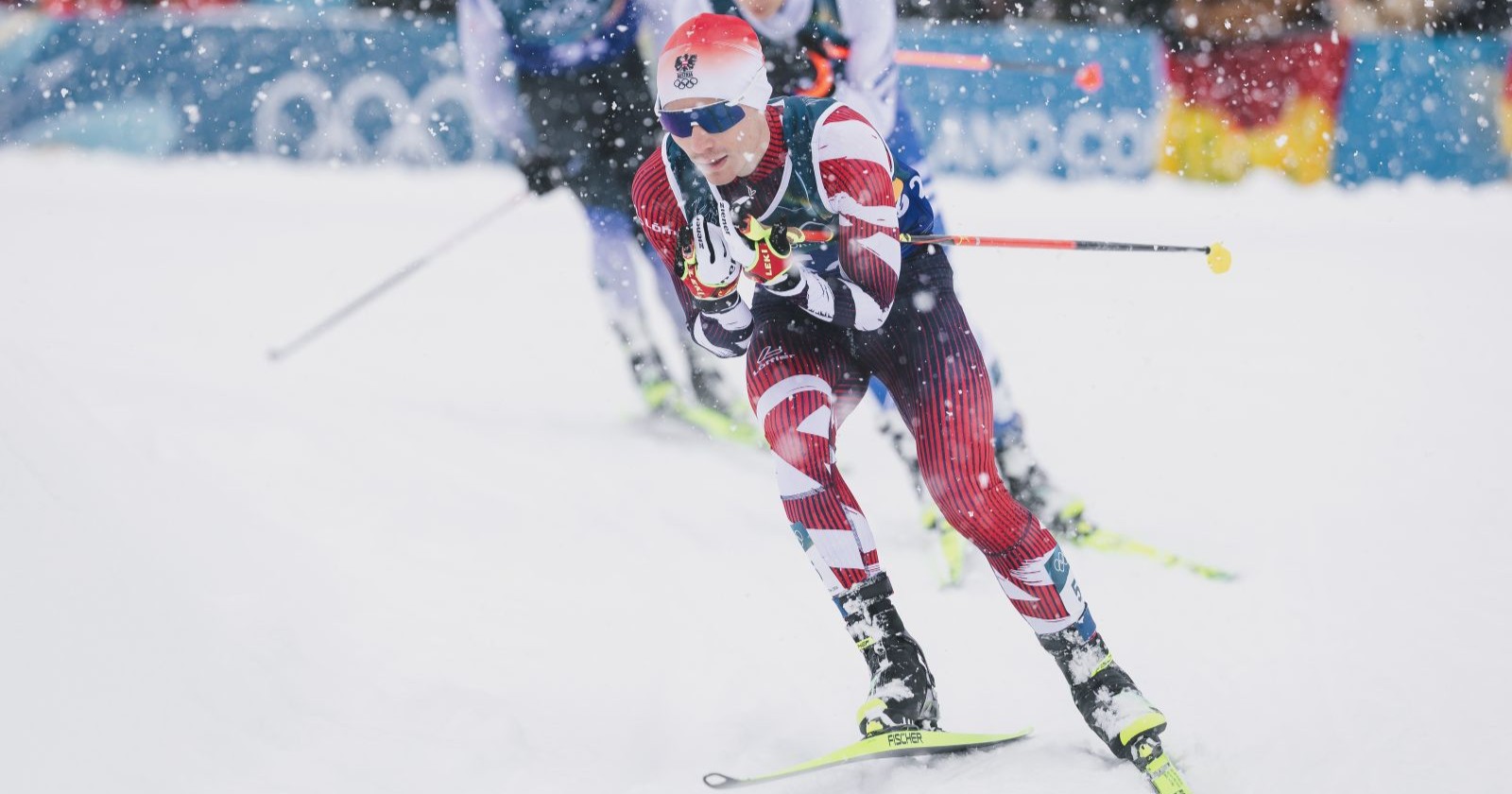 Ein österreichischer Nordischer Kombinierer in rot-weißem Rennanzug läuft bei starkem Schneefall in der Loipe während des olympischen Teamsprints in Val di Fiemme.