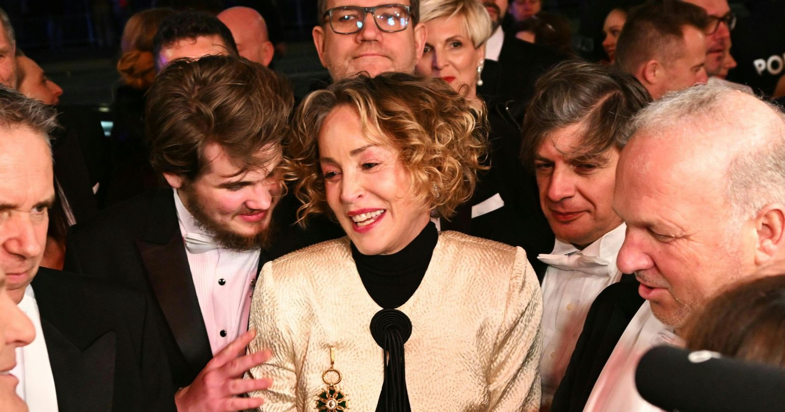 Sharon Stone in goldenem Kleid beim Wiener Opernball, lächelnd und umringt von Ballgästen, während sie mit sichtlich gerührter Miene Gespräche führt.