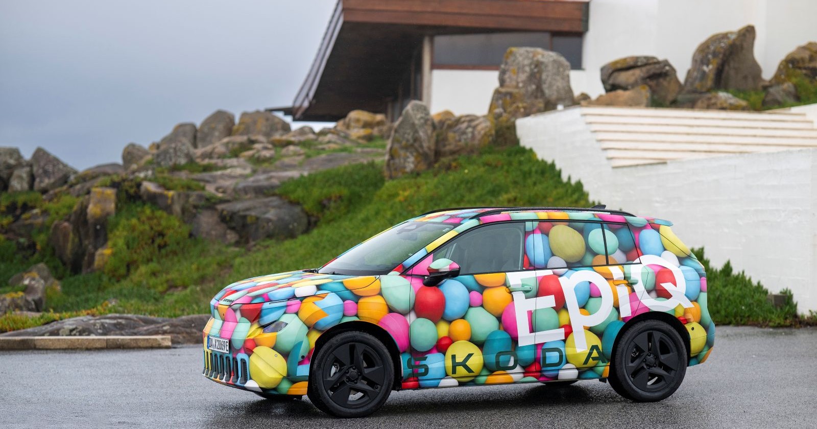 Ein Skoda Epiq steht vor einem grünen Hügel auf dem ein Haus und Felsen zu sehen sind. Das Auto hat keine einheitliche Farbe, vielmehr sind runde "Bälle" in verschiedensten Farben aufgetragen.