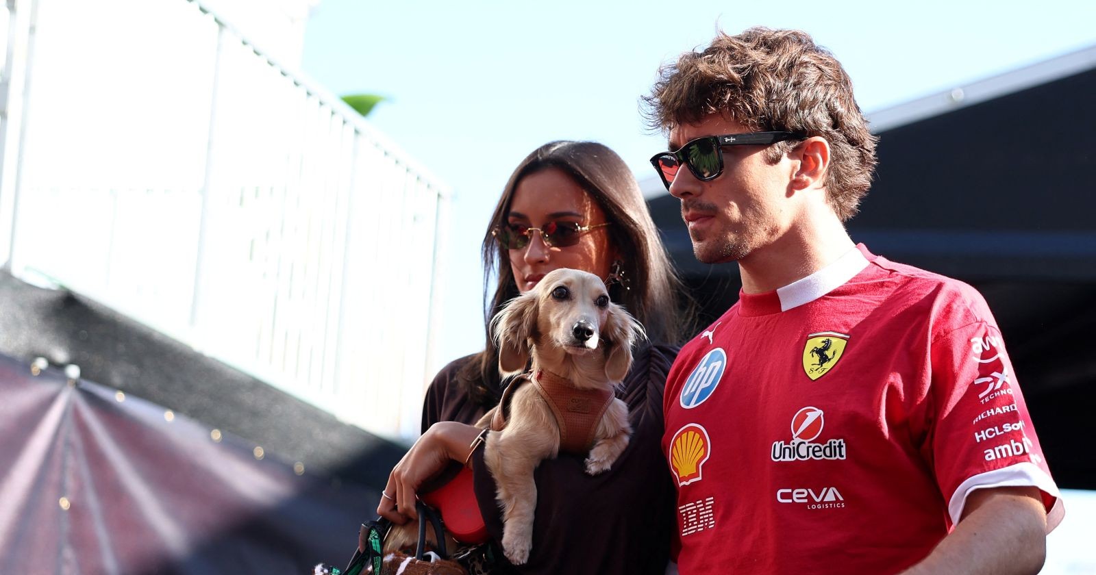 Charles Leclerc in Ferrari-Teamkleidung steht mit Alexandra Saint Mleux und Dackel Leo nebeneinander im Fahrerlager.