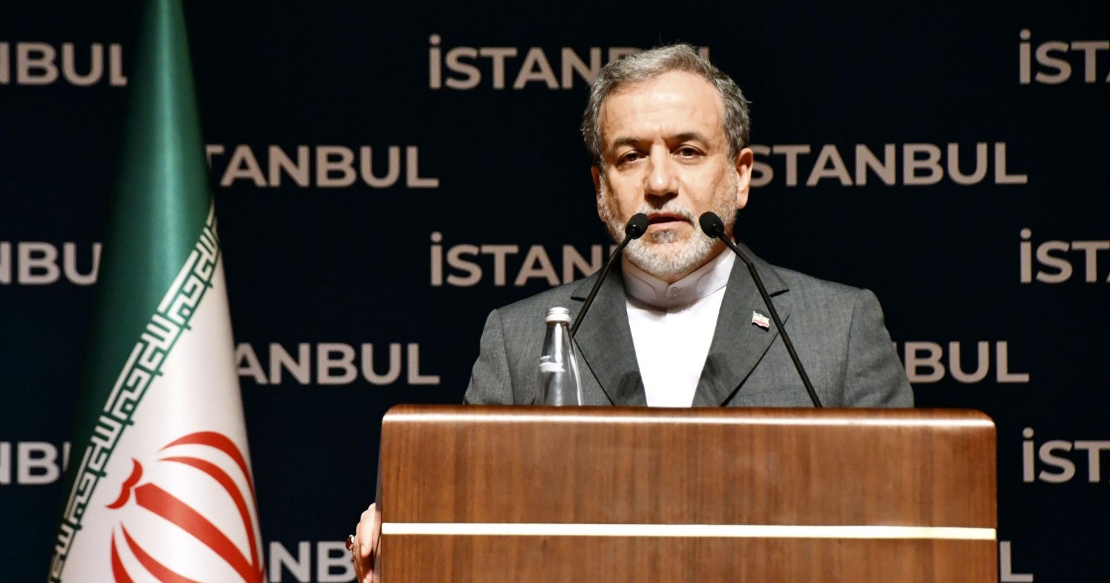 Iranischer Politiker steht am Rednerpult mit iranischer Flagge im Hintergrund während einer Veranstaltung in Istanbul.