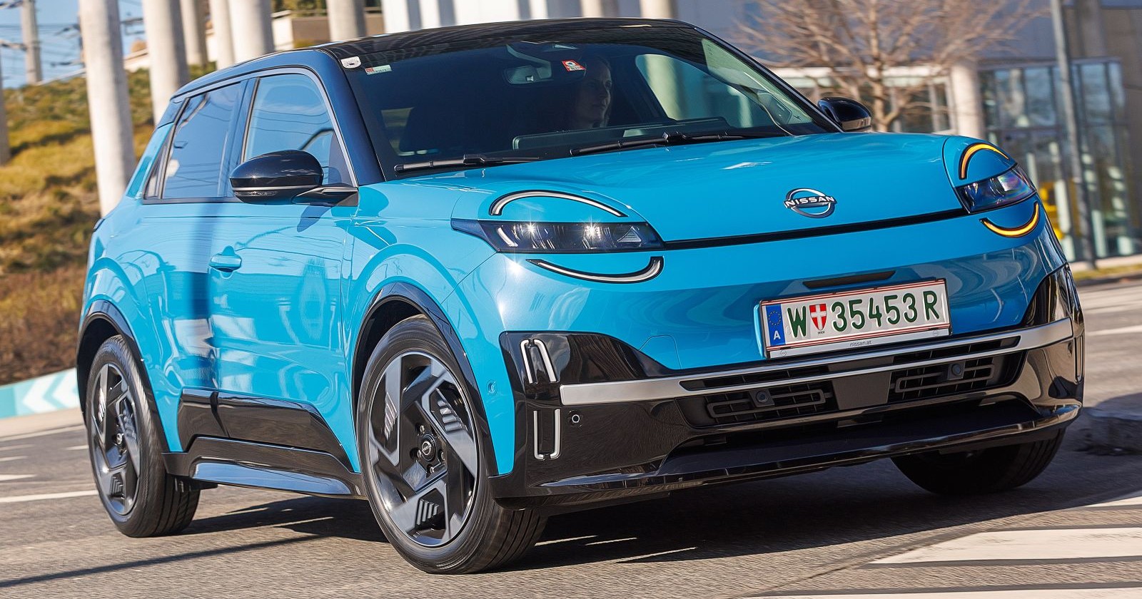 Ein hellblauer Nissan Micra glänzt in der Sonne. Das Auto parkt vor einer Glasfront, zu sehen sind auch einige Bäume.