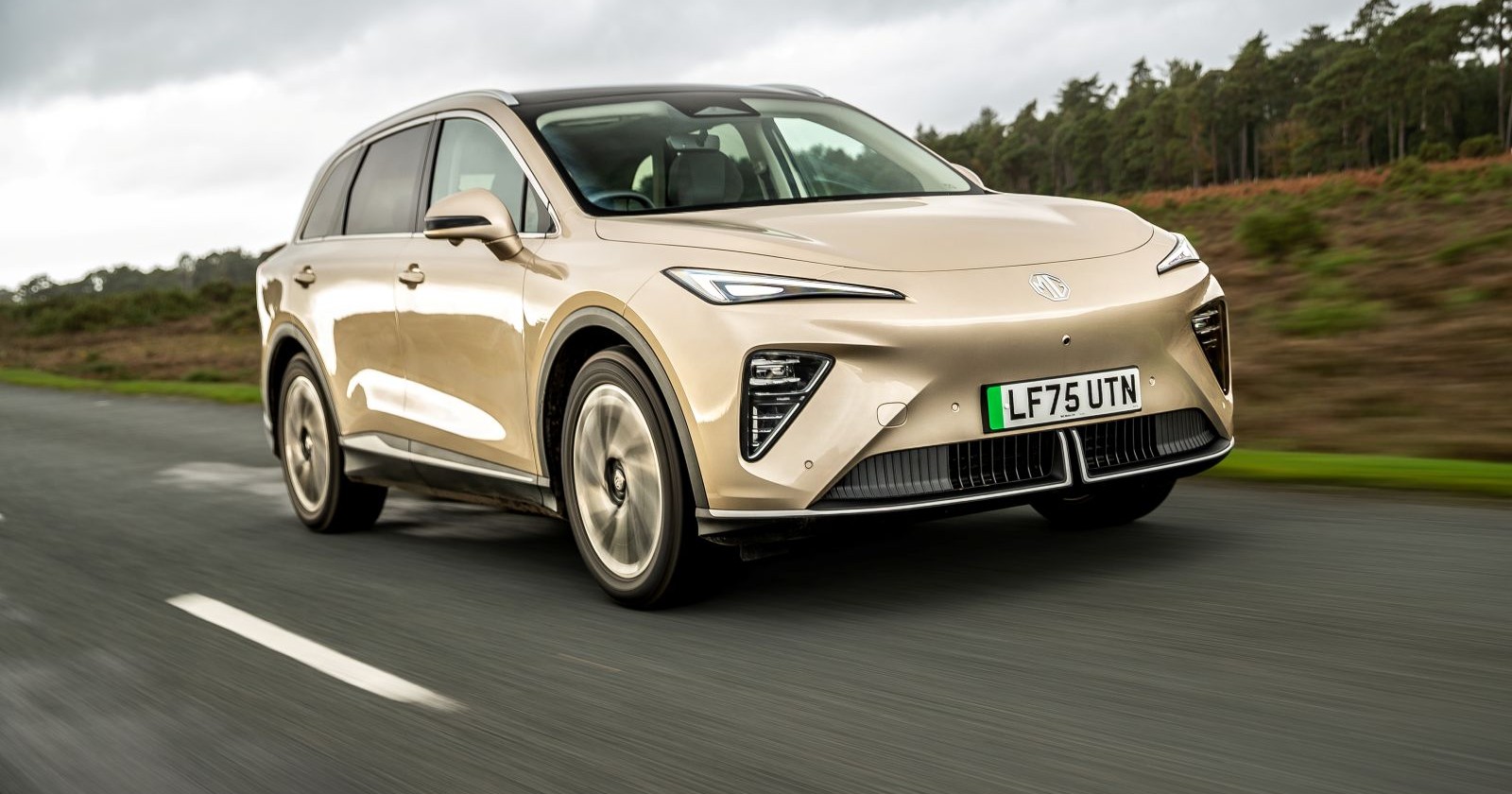 Ein hellbrauner MG MGS6, dessen Farbe an einen Milchkaffee erinnert, fährt auf einer Landstraße. Der Himmel ist bedeckt, im Hintergrund sieht man einen Laubwald.