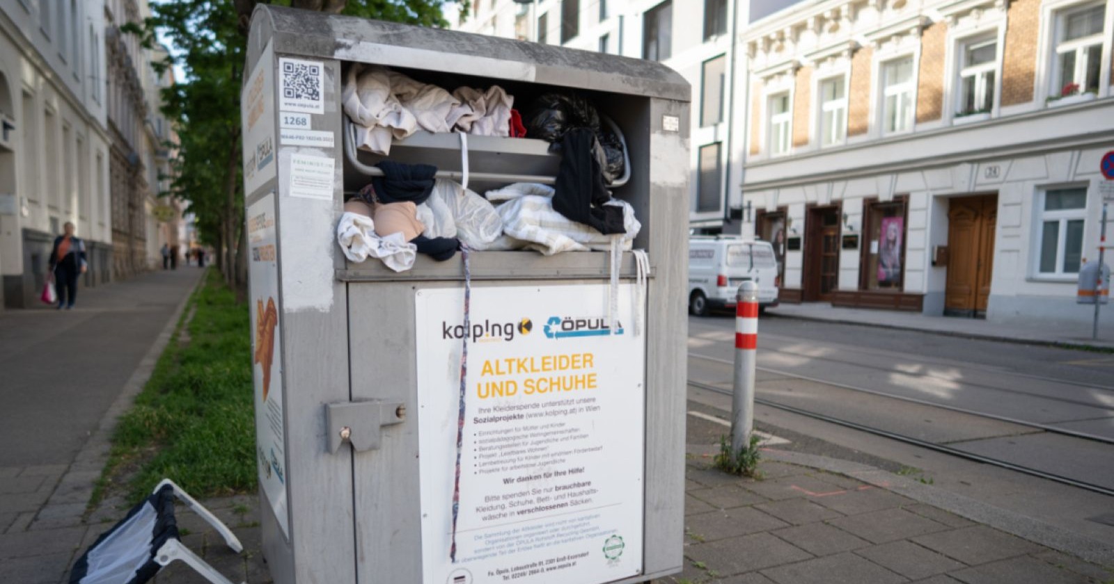 Überfüllter Altkleider-Container in Wien mit herausquellender Kleidung und danebenliegendem Müll im öffentlichen Straßenraum