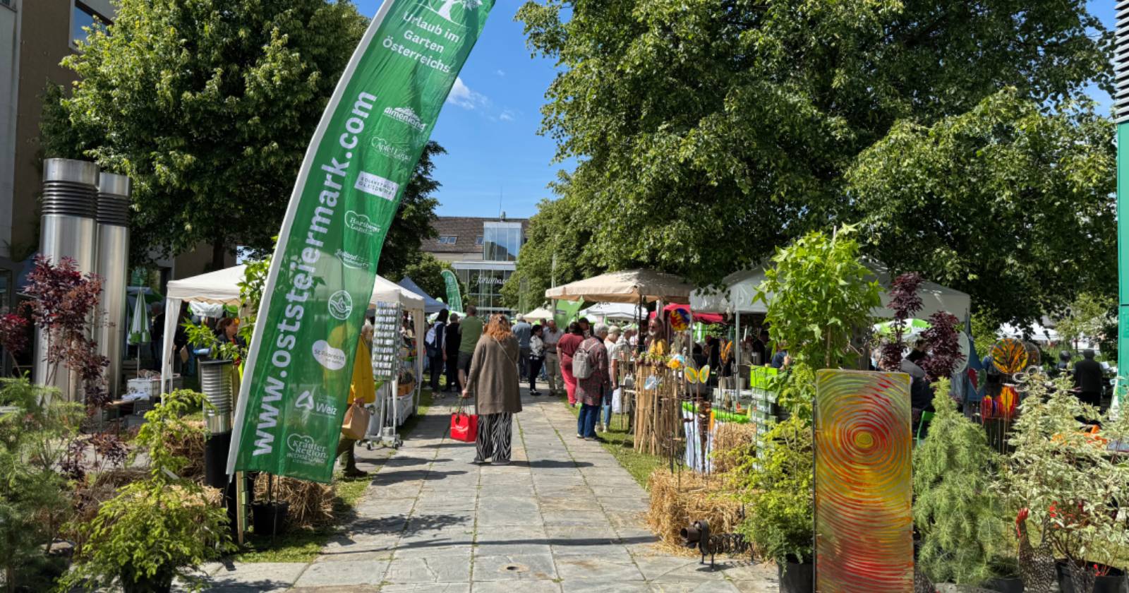 Belebter Gartenmarkt im Freien mit Ständen, Pflanzen und Dekoration. Menschen spazieren zwischen den Verkaufsständen, daneben steht eine große grüne Fahne mit „oststeiermark.com“ | Credit: Stadtmarketing Gleisdorf