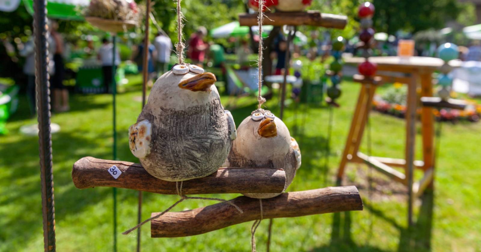 Zwei handgefertigte Ton-Vögel hängen an Schnüren auf Holzstäben. Im Hintergrund sieht man unscharf ein Gartenfest mit Menschen und Ständen | Credit: Gernot Muhr