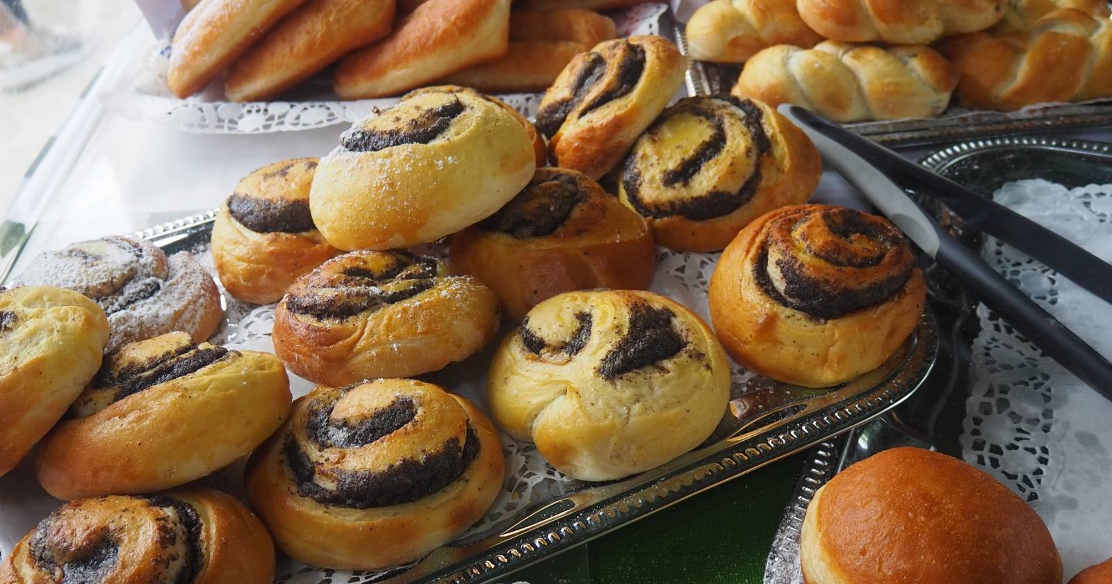 Mehrere süße Gebäckstücke (vor allem Mohnschnecken) liegen auf einem Tablett. Im Hintergrund sieht man weiteres Gebäck wie Brötchen und geflochtene Backwaren | Credit: Stadtmarketing Gleisdorf