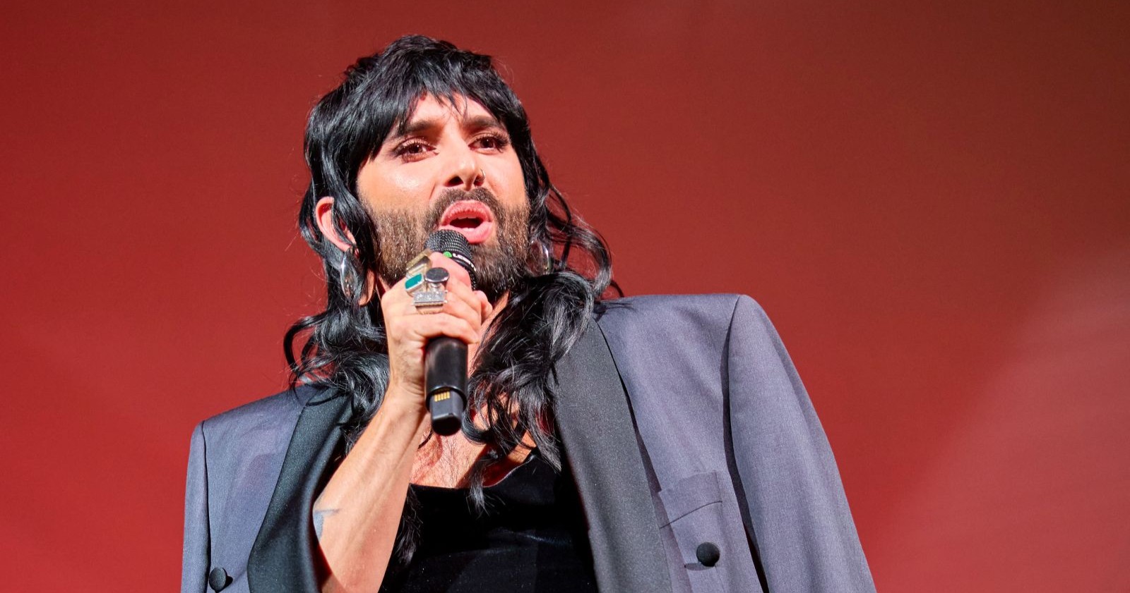 Conchita Wurst singt auf der Bühne beim Eurovision Song Contest mit Mikrofon in der Hand vor rotem Hintergrund