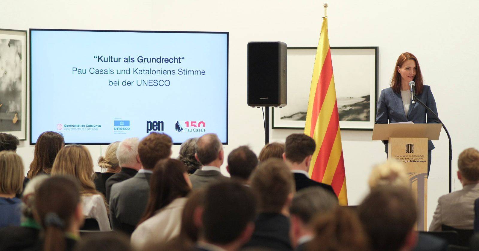 Frau im Hosenanzug spricht am Podium zu den Gästen im Publikum