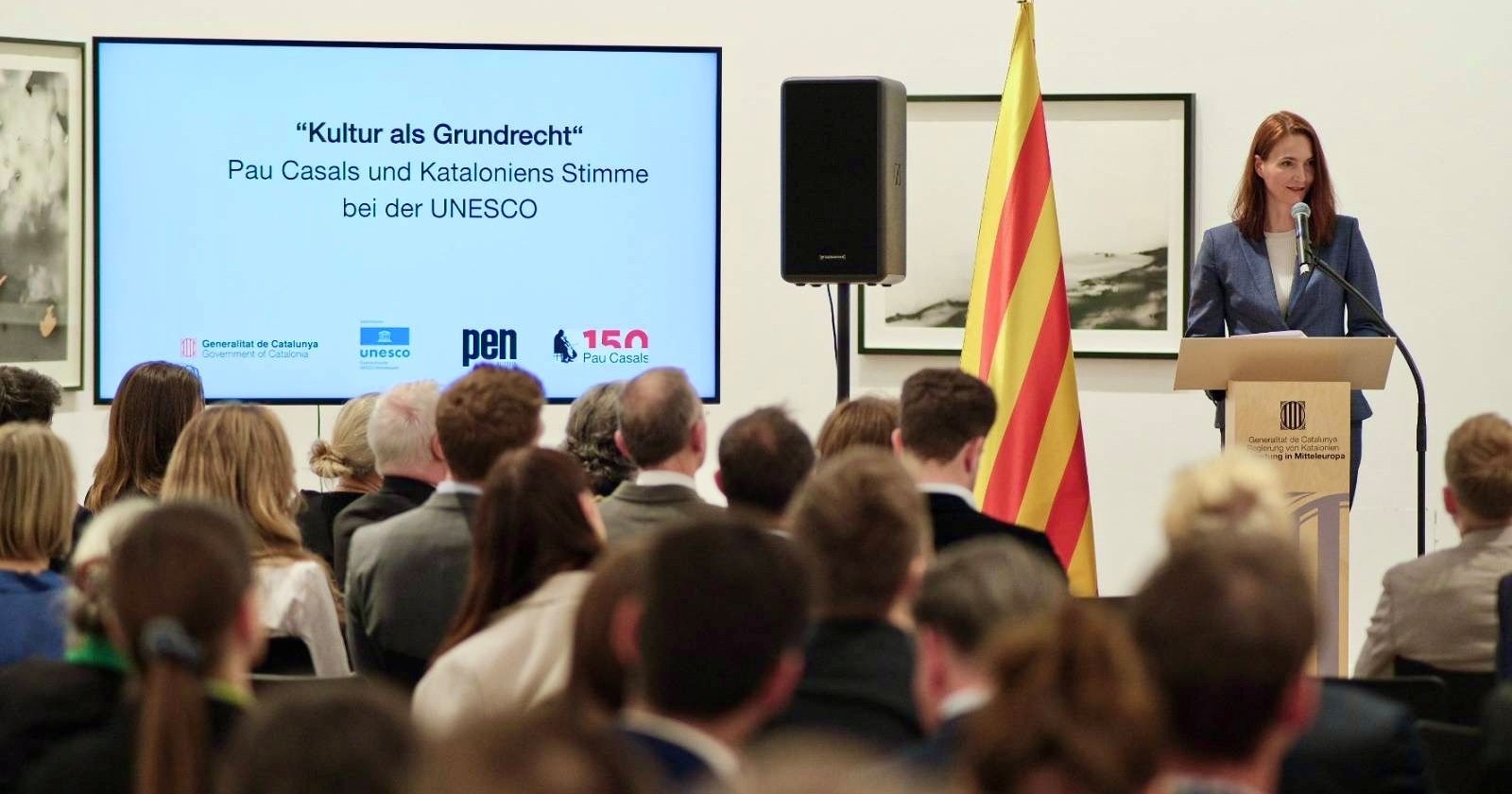 Frau im Hosenanzug spricht am Podium zu den Gästen im Publikum