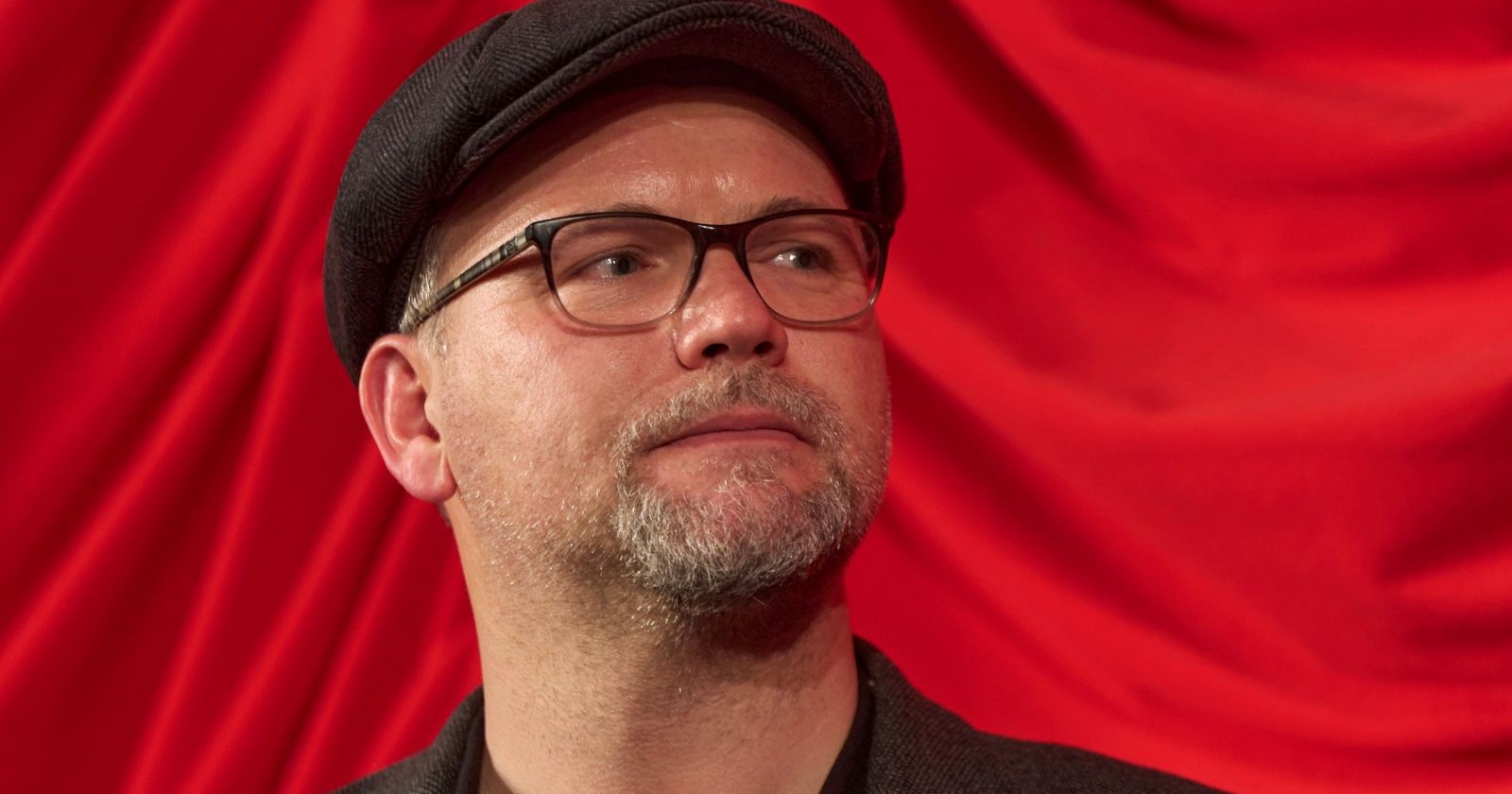 Thomas Stipsits mit Brille und Mütze vor rotem Hintergrund nach Absage seiner Auftritte wegen Schicksalsschlag