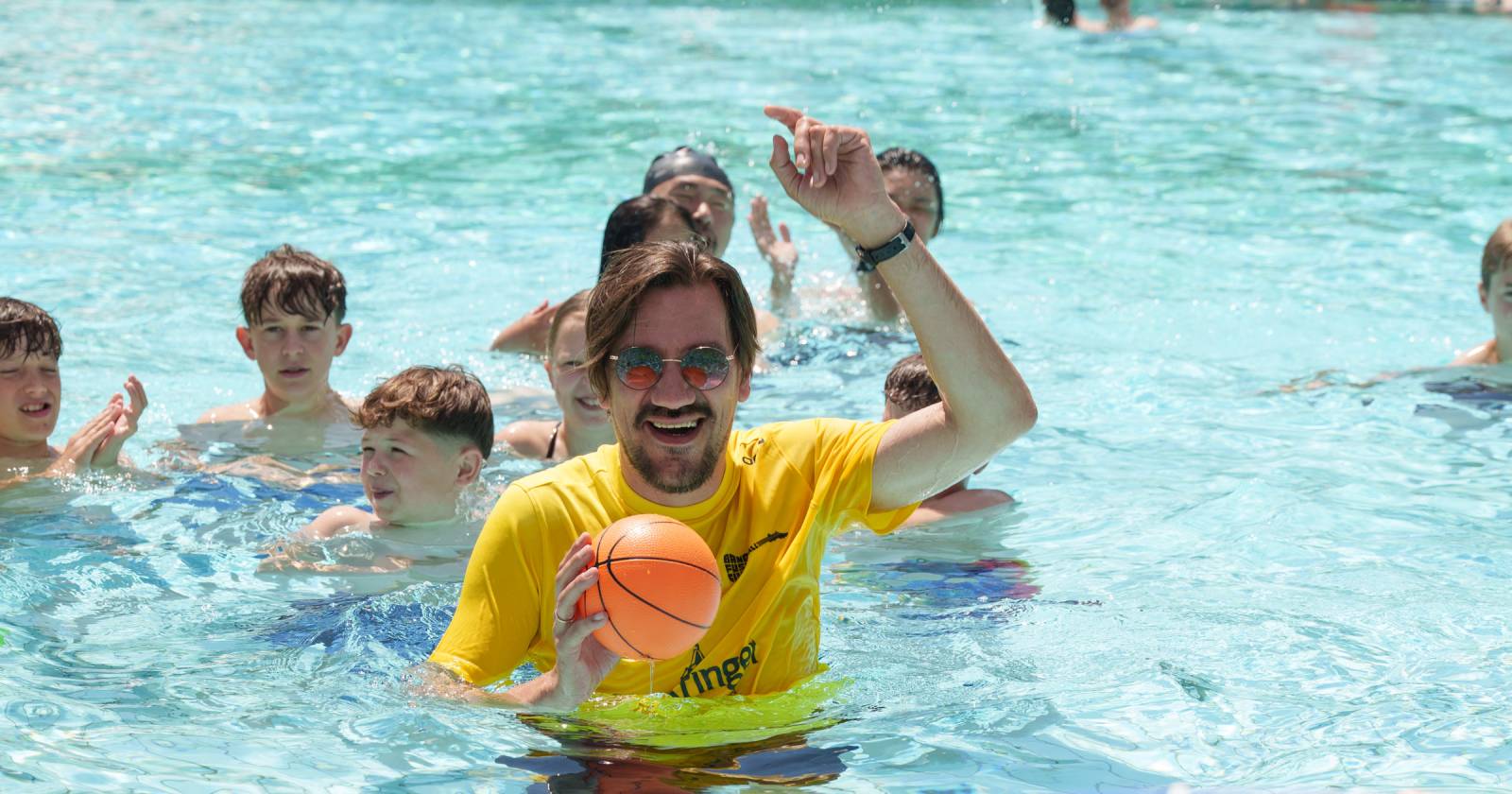 Abbildung einer Menschenmenge im Pool, im Vordergrund: ein Mann mit einem Ball in der Hand