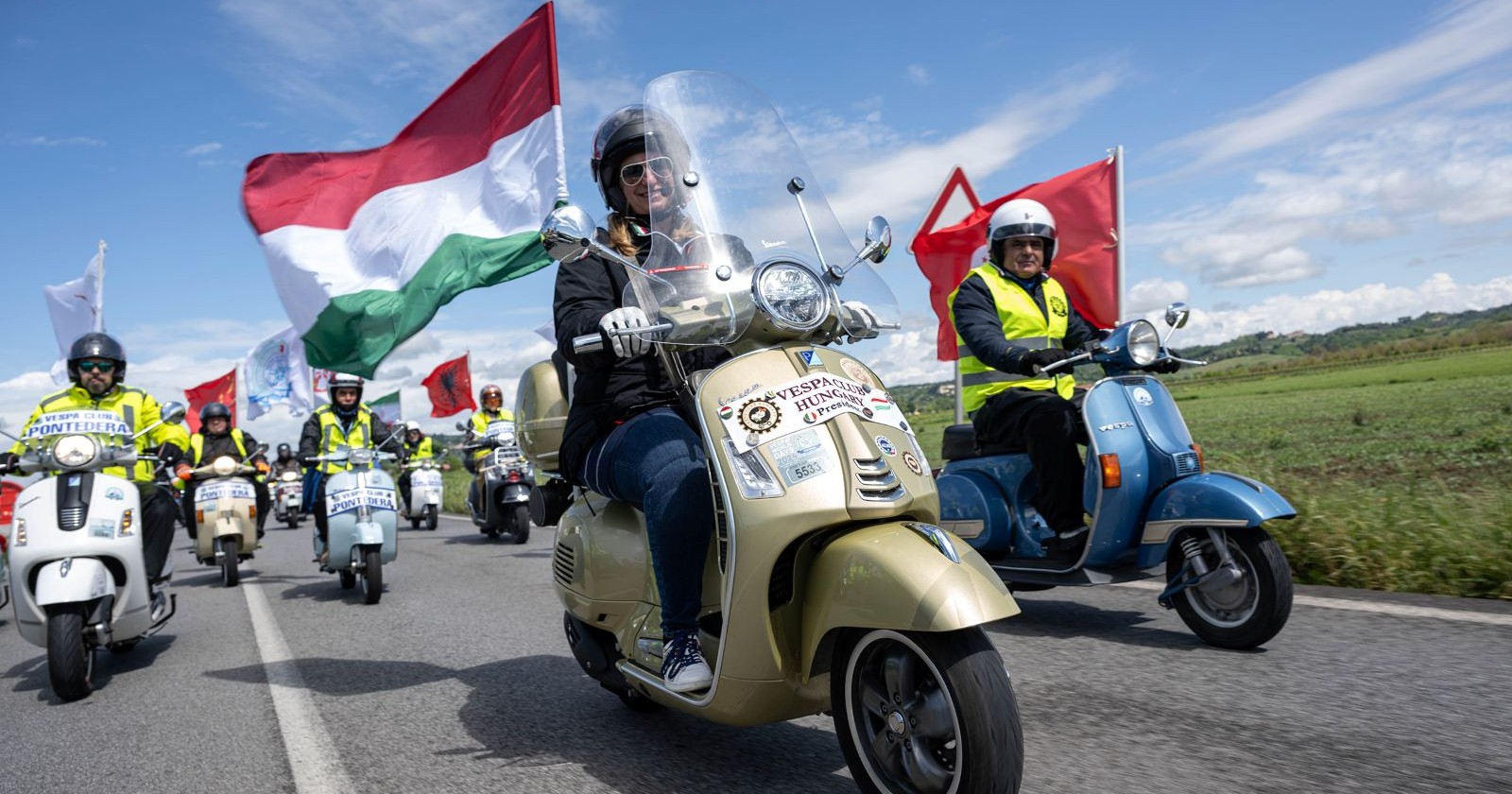 Eine Gruppe von Vespa-Fahrern auf weißen, blauen und goldenen Vespas fahren auf einer Landstraße. Alle tragen einen Helm, manche fahren mit der italienischen Flagge.