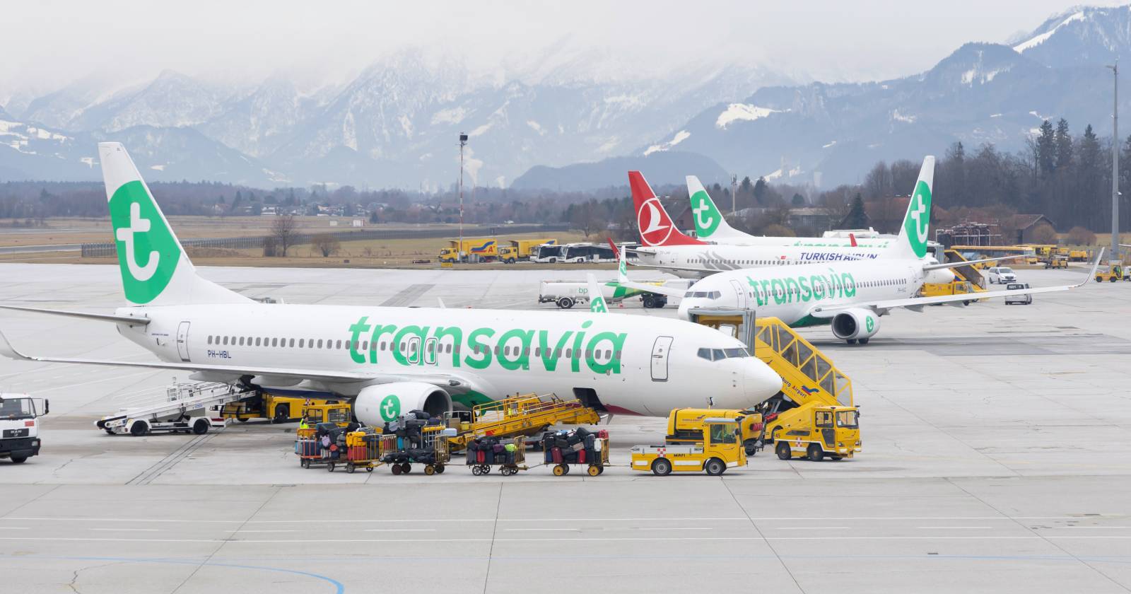 Mehrere Flugzeuge der Airline Transavia stehen am Salzburg Airport auf dem Vorfeld, während Gepäck verladen und Passagiere abgefertigt werden. Im Hintergrund sind weitere Maschinen sowie die verschneite Bergkulisse zu sehen.
