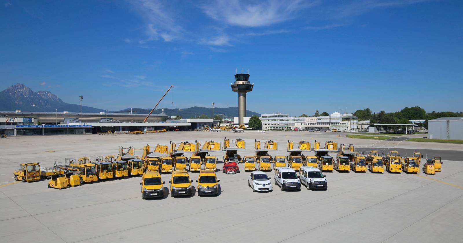Am Salzburg Airport sind zahlreiche gelbe Flughafenfahrzeuge und Geräte ordentlich auf dem Vorfeld aufgestellt. Im Hintergrund sind der Tower, das Flughafengebäude sowie Berge unter strahlend blauem Himmel zu sehen.