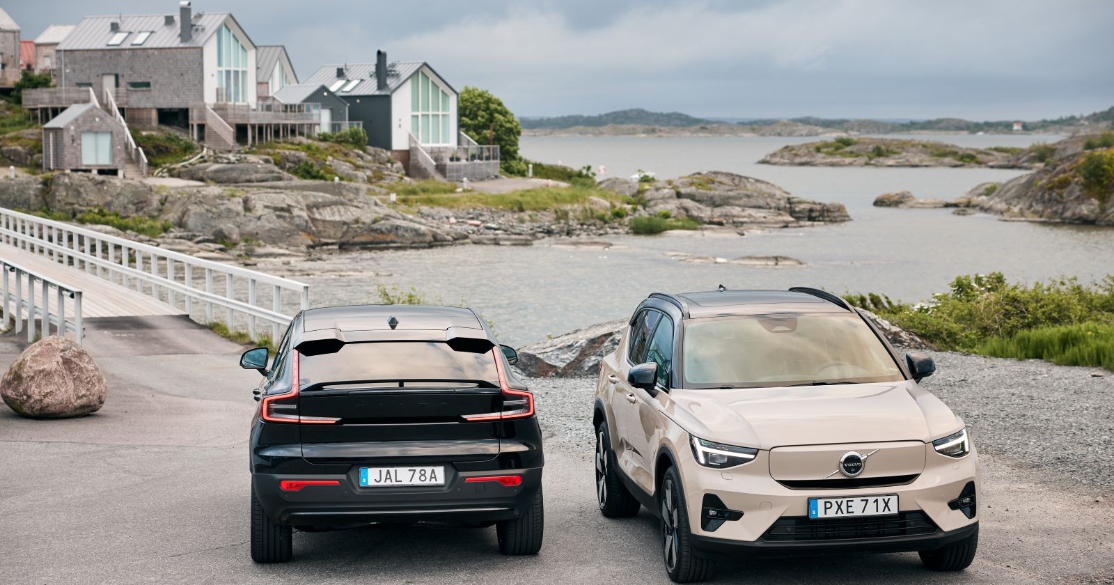 Ein schwarzer Volvo EC40 und ein hellbrauner Volvo EX40 parken am Rande einer Meeresbucht. Im Hintergrund sind einige Häuser zu sehen, am Himmel einige Wolken.