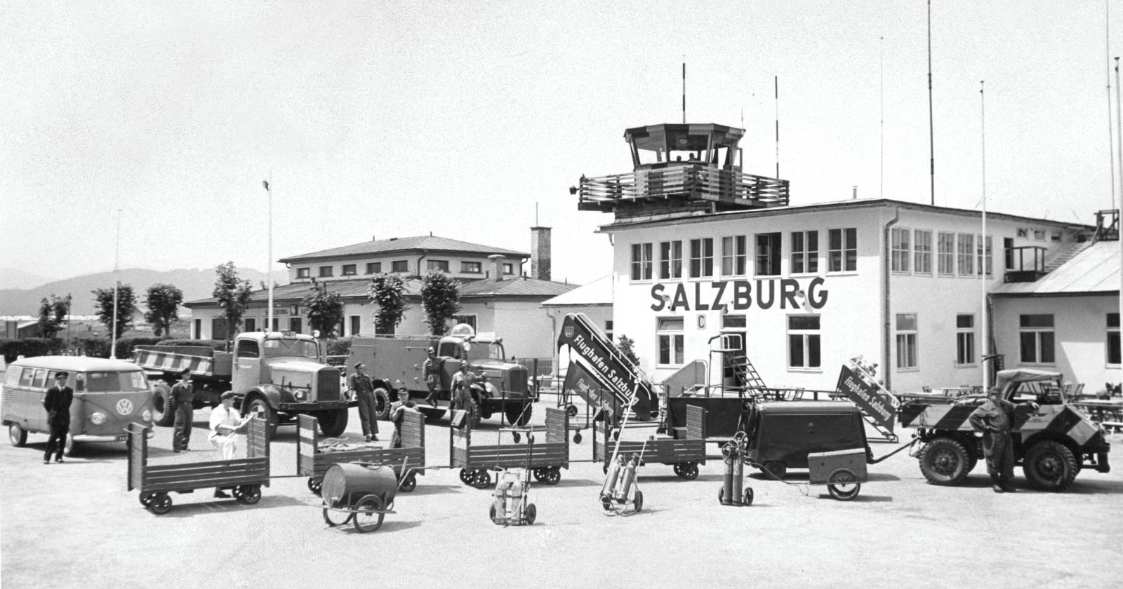 Historische Schwarz-Weiß-Aufnahme des Flughafens Salzburg aus dem Jahr 1956 mit alten Fahrzeugen, Gepäckwagen und Flughafenmitarbeitern vor dem damaligen Terminalgebäude und Kontrollturm.