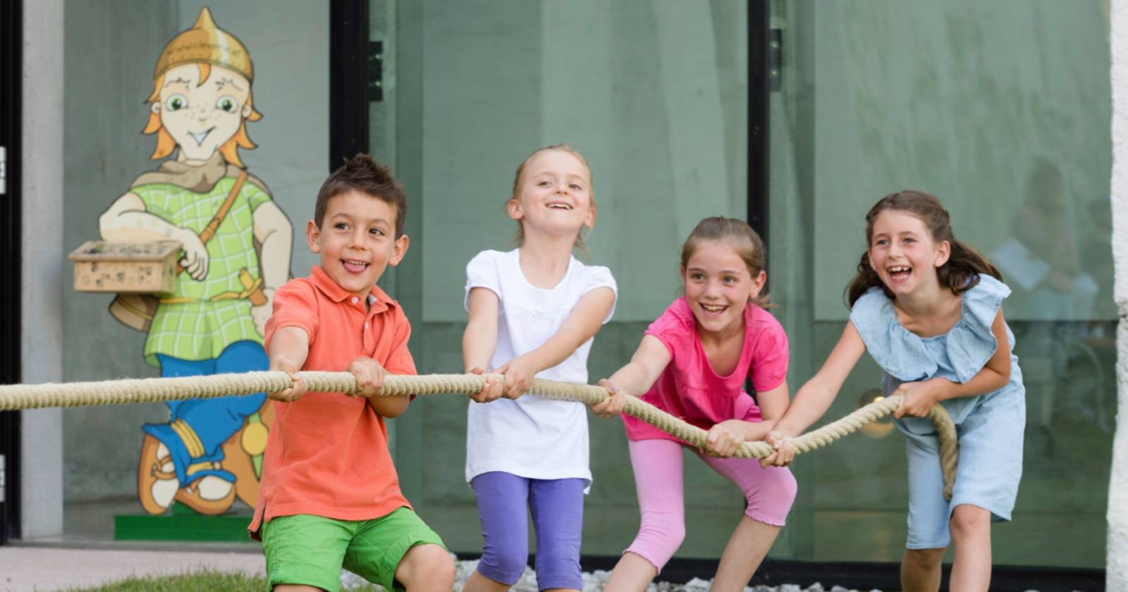 Vier Kinder ziehen gemeinsam lachend an einem Seil und spielen ausgelassen im Freien. Im Hintergrund ist eine kindgerechte Figur vor einem Gebäude zu sehen | Credit: Bryan Reinhart
