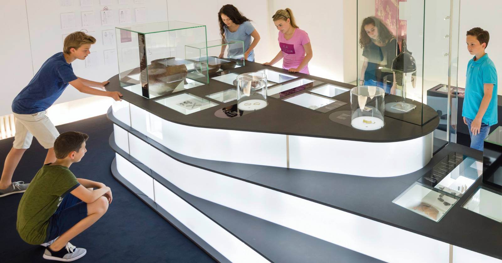 Mehrere Kinder erkunden interessiert eine moderne Museumsausstellung mit interaktiven Vitrinen und historischen Exponaten. Die Jugendlichen beobachten und entdecken die ausgestellten Objekte gemeinsam | Credit: Bryan Reinhart