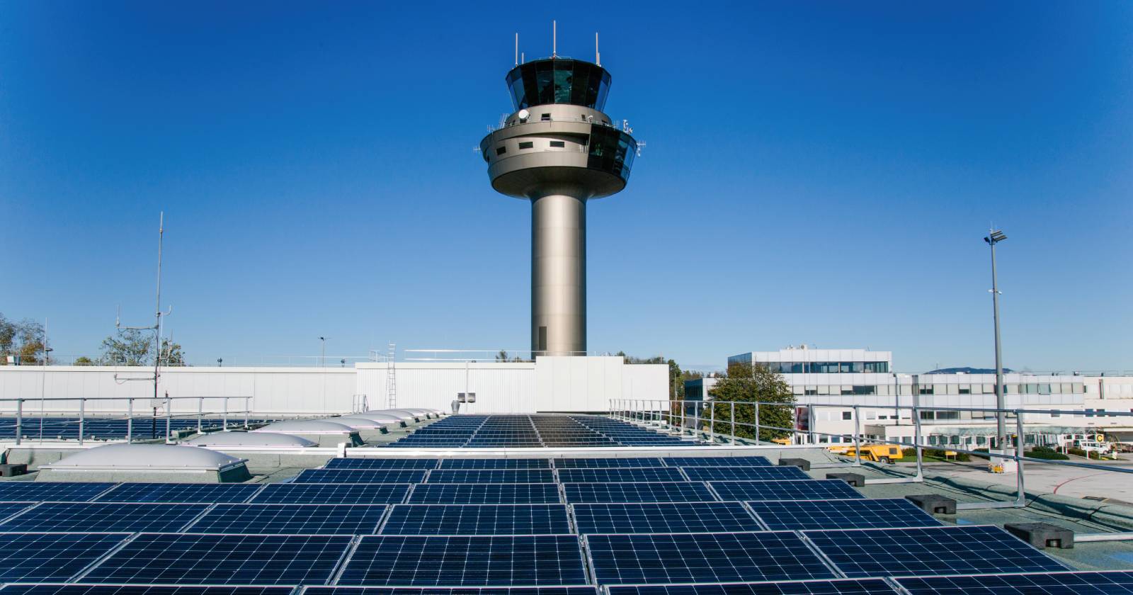 Photovoltaikanlagen auf dem Dach des Salzburg Airport vor dem modernen Towergebäude unter klarem blauem Himmel. Die Solarpaneele stehen für nachhaltige Energieversorgung am Flughafen.