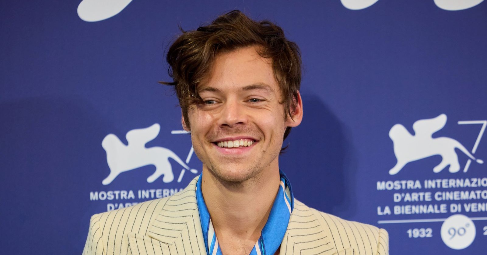 Harry Styles lächelt bei einem öffentlichen Auftritt in hellem Nadelstreifen-Anzug vor blauem Event-Hintergrund.