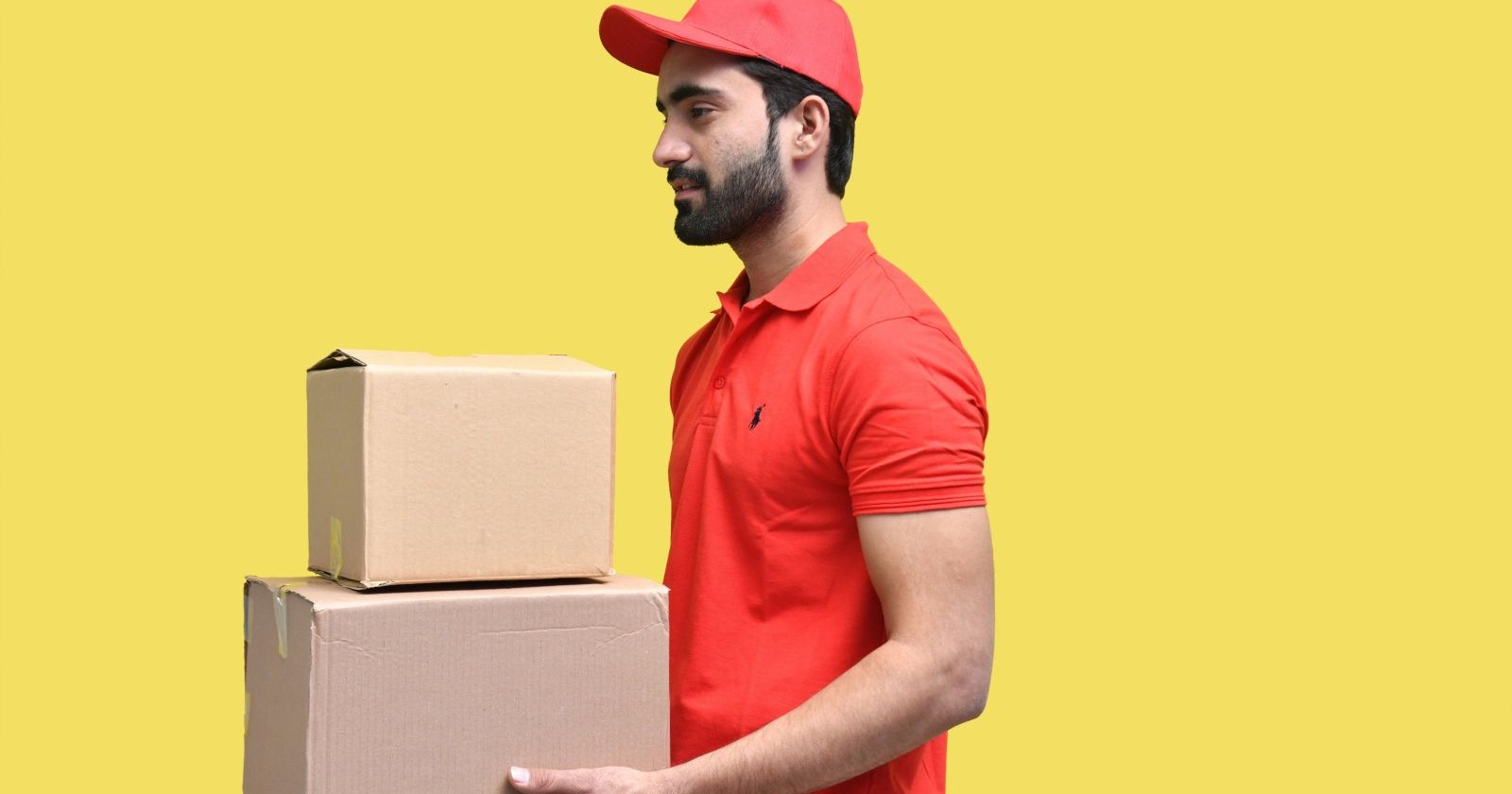 Paketzusteller mit rotem Polo und rotem Kapperl mit zwei Paketen vor einem gelben Hintergrund