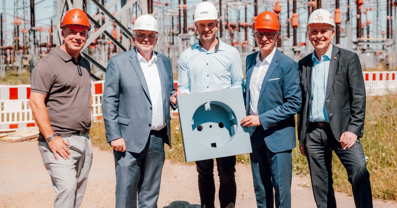 Fünf Männer in Bürokleidung mit Baustellenhelmen an einer sonnigen Baustelle