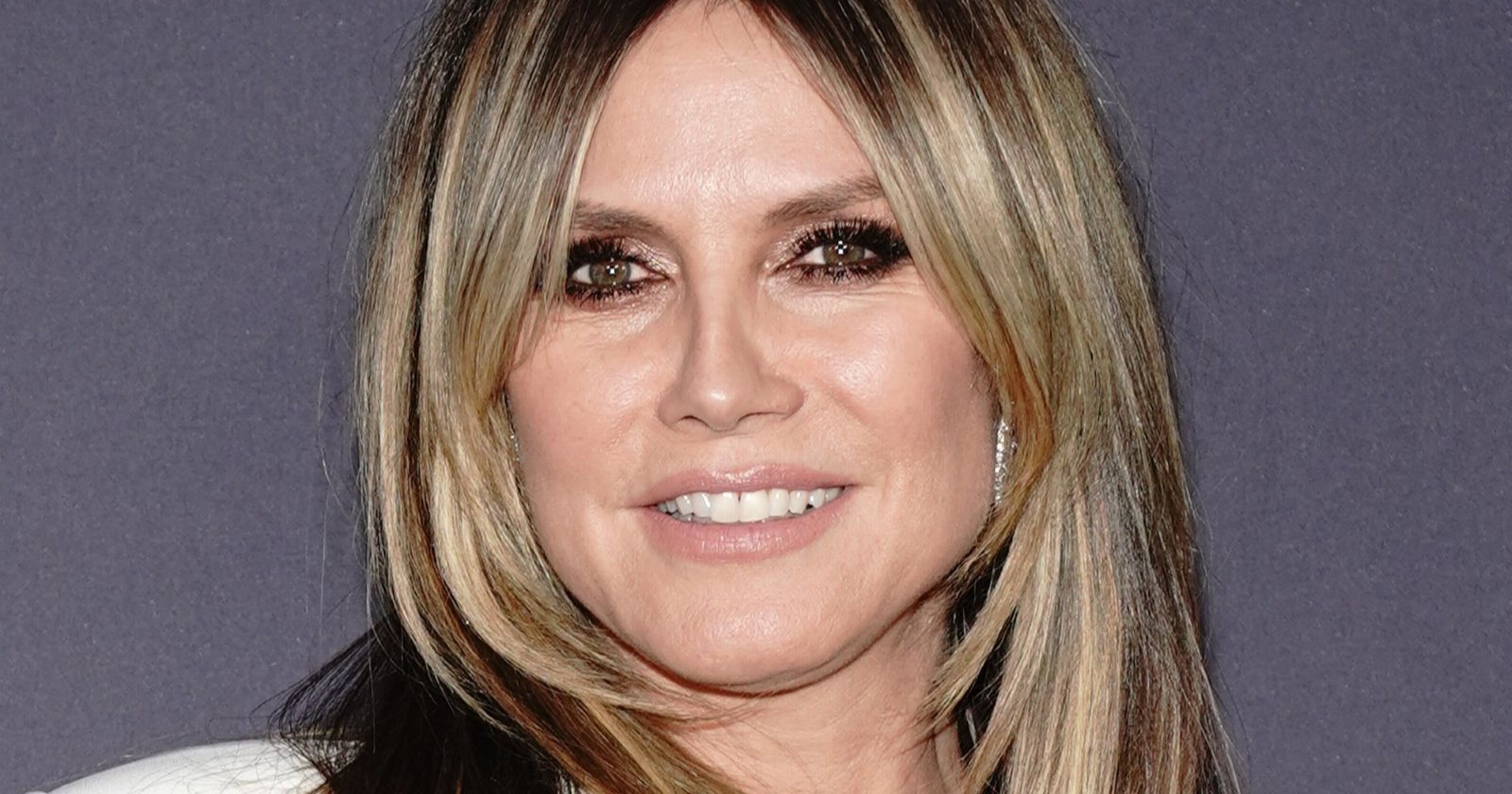 Heidi Klum lächelt bei einem öffentlichen Auftritt in die Kamera. Das Topmodel trägt ein weißes Outfit und ihre blonden Haare mit dunklem Ansatz offen.