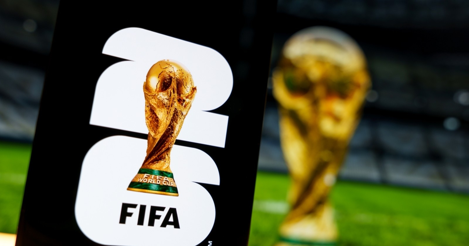 Offizielles FIFA World Cup 2026 Markenlogo auf einem digitalen Bildschirm neben einem unscharfen Trophäenhintergrund.