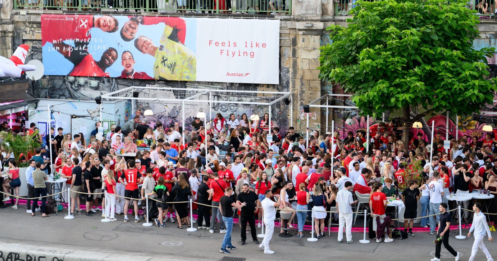 Menschenmenge beim Public Viewing am Donaukanal in Wien während eines Fußballspiels, viele Fans in rot-weiß gekleidet, Szene vor der Eskalation der Gewalt