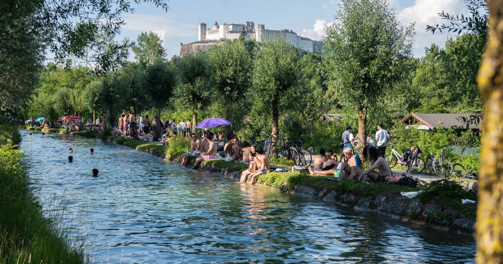 Viele Menschen baden im Almkanal in Salzburg an einem Sommertag, im Hintergrund ist die Festung Hohensalzburg zu sehen; Gülle nach einem Unwetter verursacht Erkrankungen bei Badegästen