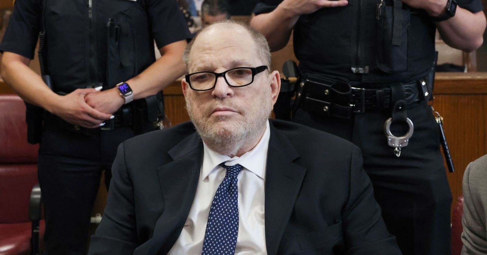 Harvey Weinstein sitzt während eines Gerichtstermins im Anzug vor Polizisten. Der Ex-Hollywood-Produzent wurde nach einem Herzversagen aus dem Gefängnis ins Krankenhaus gebracht.