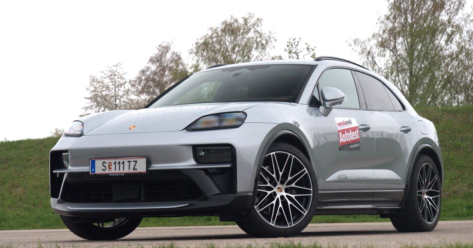 Ein silberner Porsche Macan GTS parkt vor einer Böschung. Im Hintergrund sind einige Laubbäume zu sehen.