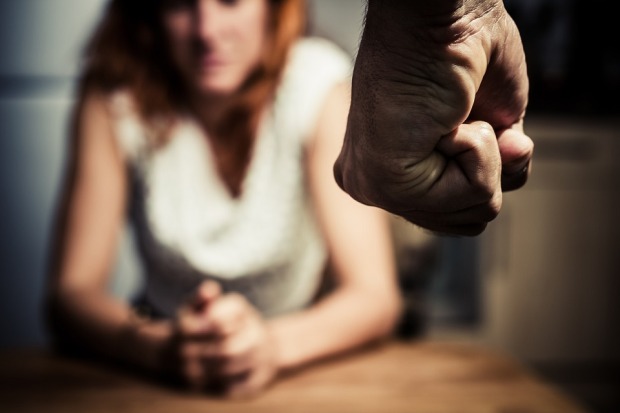 Unscharf im Hintergrund sieht man eine Frau sitzen. Sie hat die Hände gefaltet. Im Vordergrund eine geballte Männerfaust.