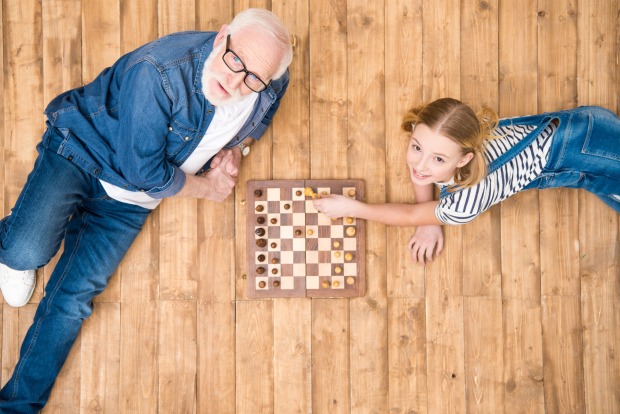 Ein älterer Herr spielt mit einem jungen Mädchen auf einem Holzboden liegend Schach