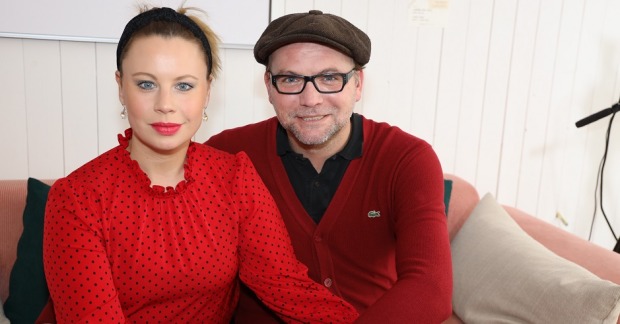 Katharina Straßer und Ehemann Thomas Stipsits sitzen Arm in Arm auf einer Couch. Sie schauen direkt in die Kamera.