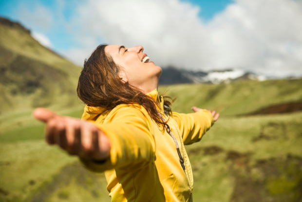 Eine Frau in einem gelben Regenmantel blickt in Island lachend und mit weit ausgestreckten Armen zum Himmel | Credit: iStock.com/erikreis