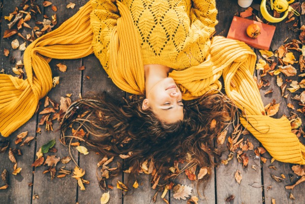 Glückliche Frau liegt an einem sonnigen Herbsttag in ihrem gelben Pullover entspannt auf der Holzterrasse | Credit: iStock.com/Maria Korneeva